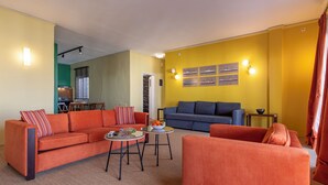 A living room with a sofa, armchairs, a coffee table, and a dining table. There is a kitchen in the background.