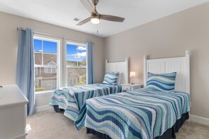 A bedroom with two beds, a ceiling fan, a window with curtains, and a nightstand with a lamp.