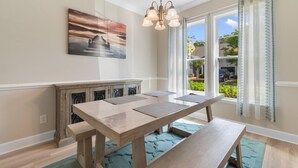 Un espace repas avec une longue table, des bancs, un lustre, une fenêtre avec des rideaux et un tableau accroché au mur.