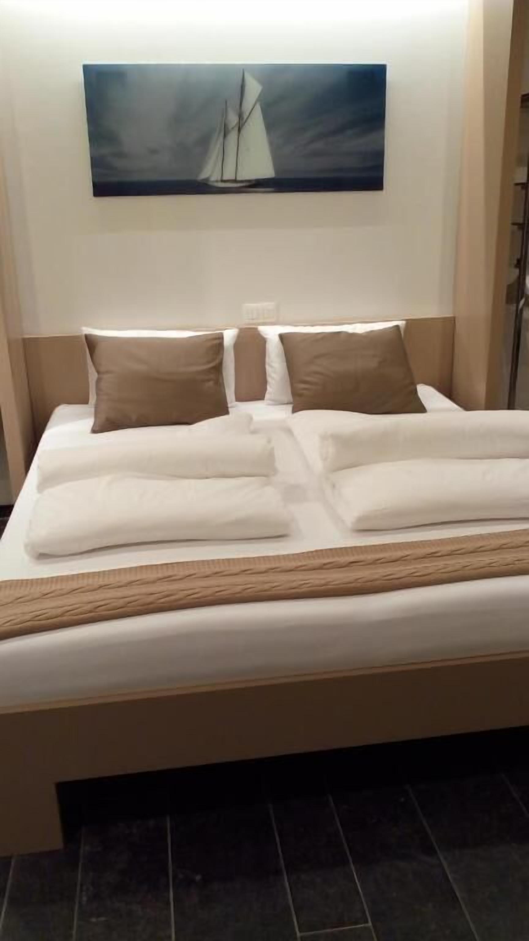Ein ordentlich bezogenes Bett mit weißen Laken und einer braunen Tagesdecke, ein Segelbootgemälde an der Wand und ein dunkler Fliesenboden.