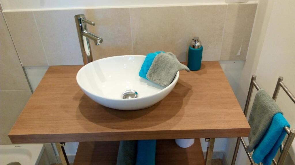 Un baño con un lavamanos blanco tipo cuenco sobre un mueble de madera, un perchero con una toalla azul y un dispensador de jabón azul.