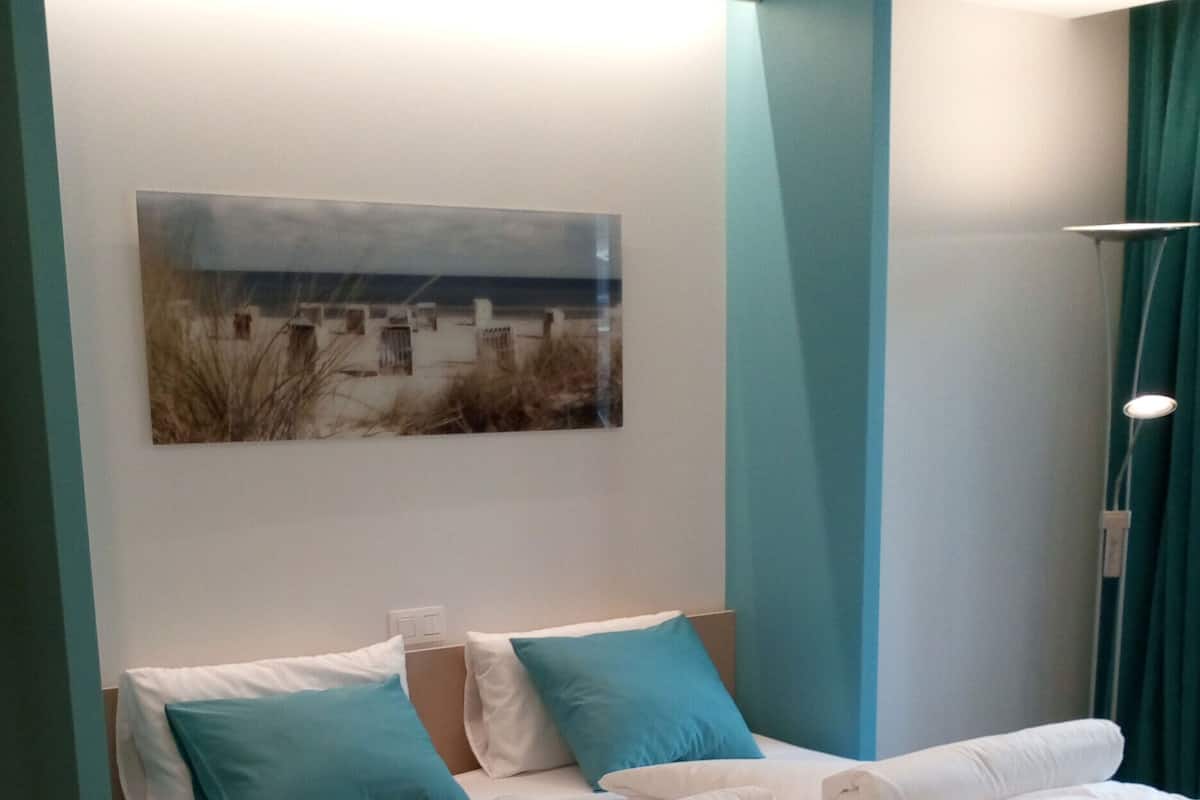 A neatly made bed with white linens and blue pillows, a painting of a beach scene on the wall, and teal-colored furniture.