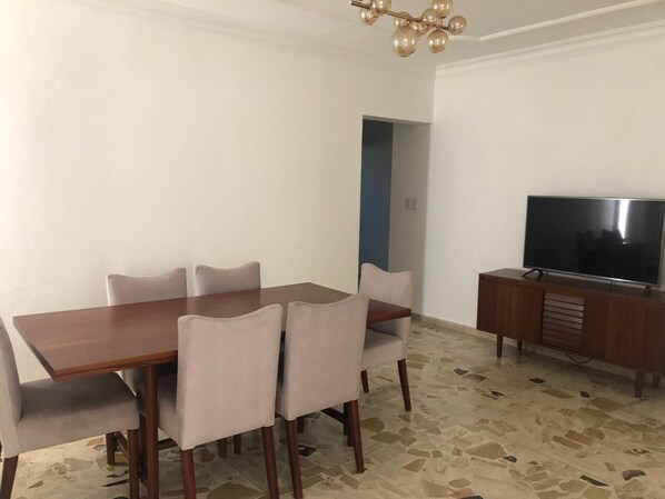 Un comedor con una mesa de madera y seis sillas, un televisor sobre un mueble de madera, y piso de baldosas.