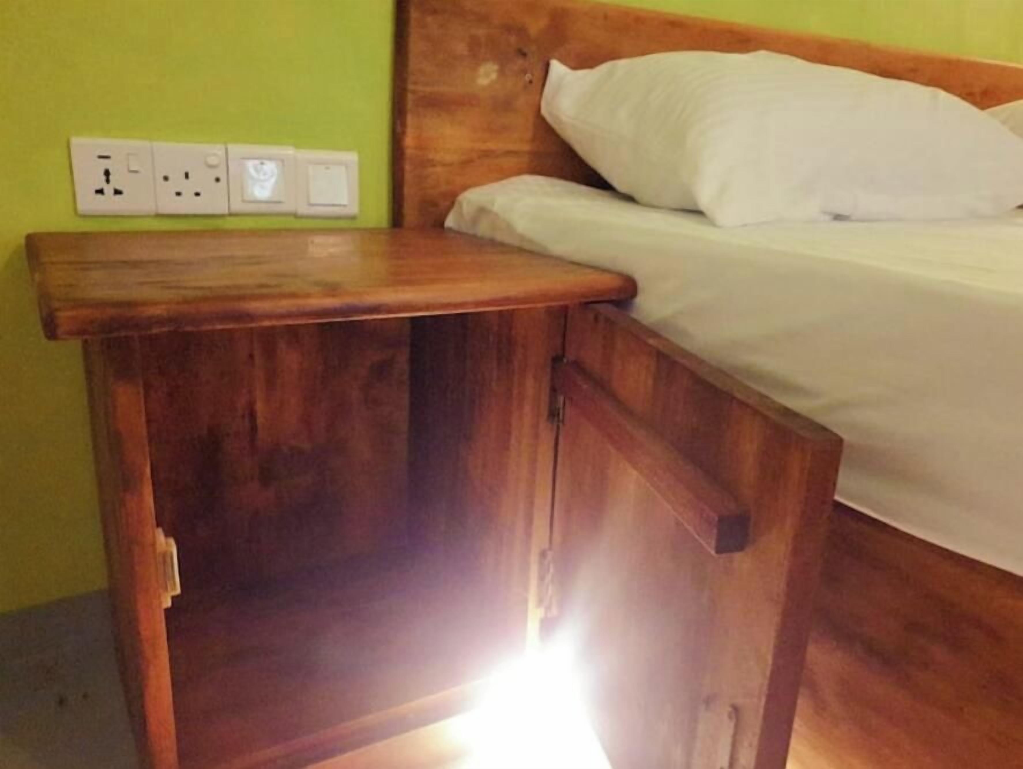 Un comodino in legno con un cassetto, un letto con biancheria bianca e una presa elettrica.