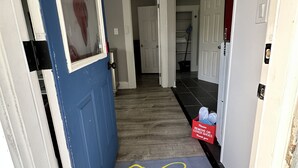 Ein Flur mit Fußmatte am Eingang, die zum Schuhe Ausziehen auffordert, eine Tür mit Schild zur Schuhentfernung und eine rote Box mit einer Botschaft zum Thema COVID-19.