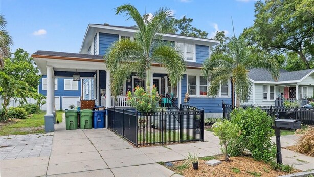 A two-story house with a front porch, a garage, and a fenced yard with palm trees.
