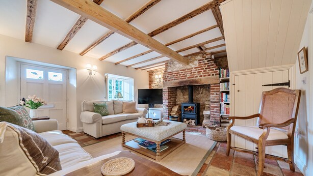 A cozy living room with exposed wooden beams, a brick fireplace, a comfortable sofa, a wooden coffee table, and a bookshelf. - Ivy Cottage - sleeps 6 guests  in 3 bedrooms (Thornham)