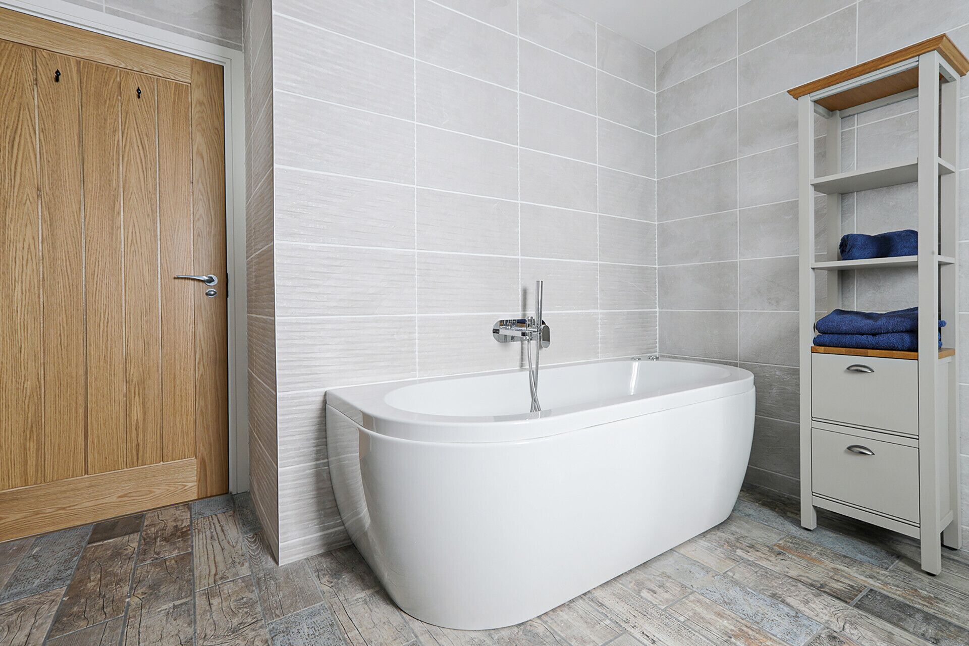 A modern bathroom with a freestanding bathtub, a wooden door, and a white tiled wall.
