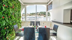 Un comedor con una mesa y sillas de vidrio, vista a edificios a través de un ventanal grande y una pared verde con plantas.