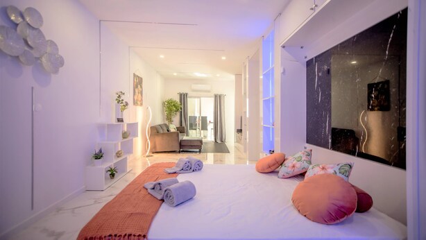 Una habitación de hotel moderna con una cama grande, una zona de estar con sofá y sillones, una mesita de noche con lámpara, y un espejo grande en la pared.