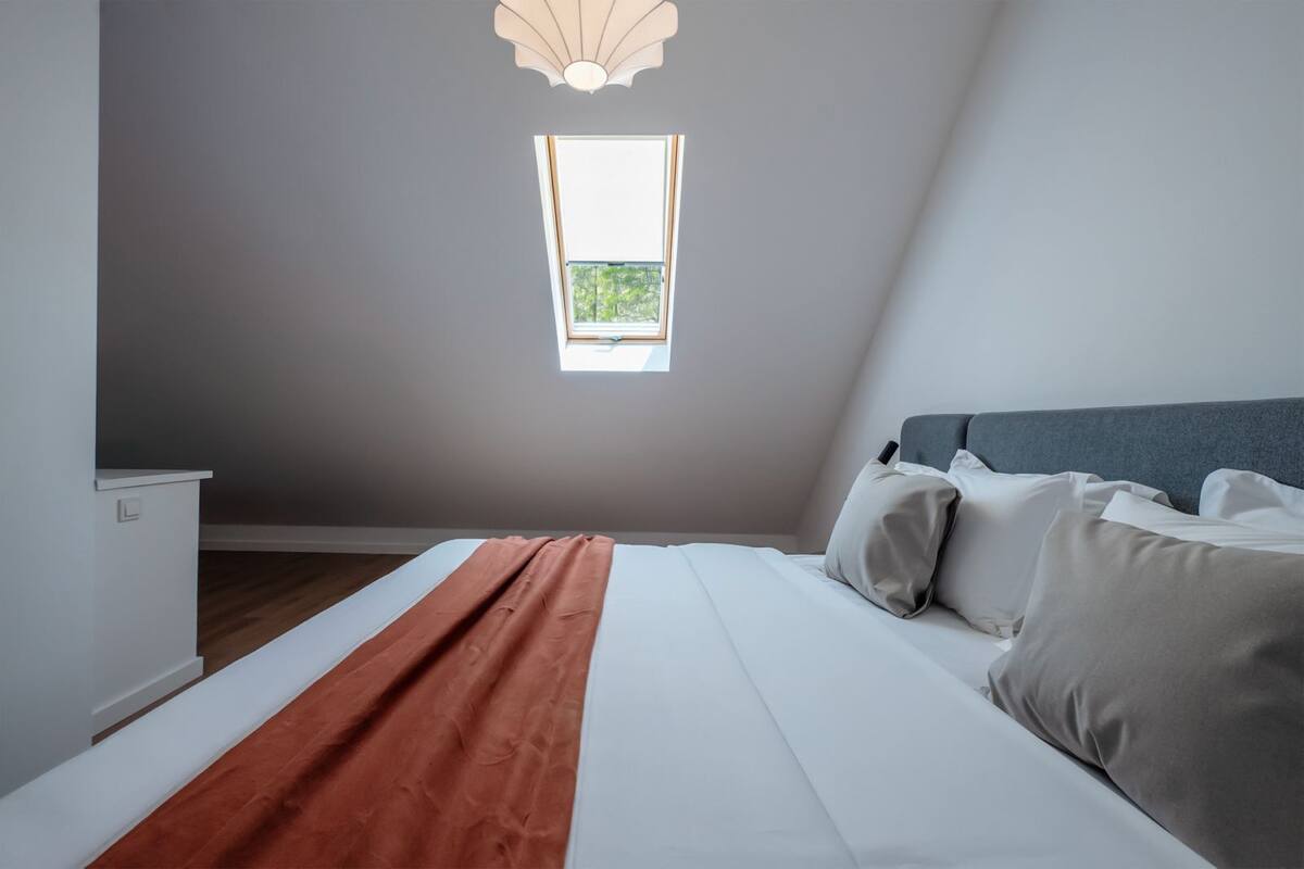 Ein Schlafzimmer mit einem Bett, Kissen, einem Nachttisch und einem Fenster mit Blick auf Grünflächen.