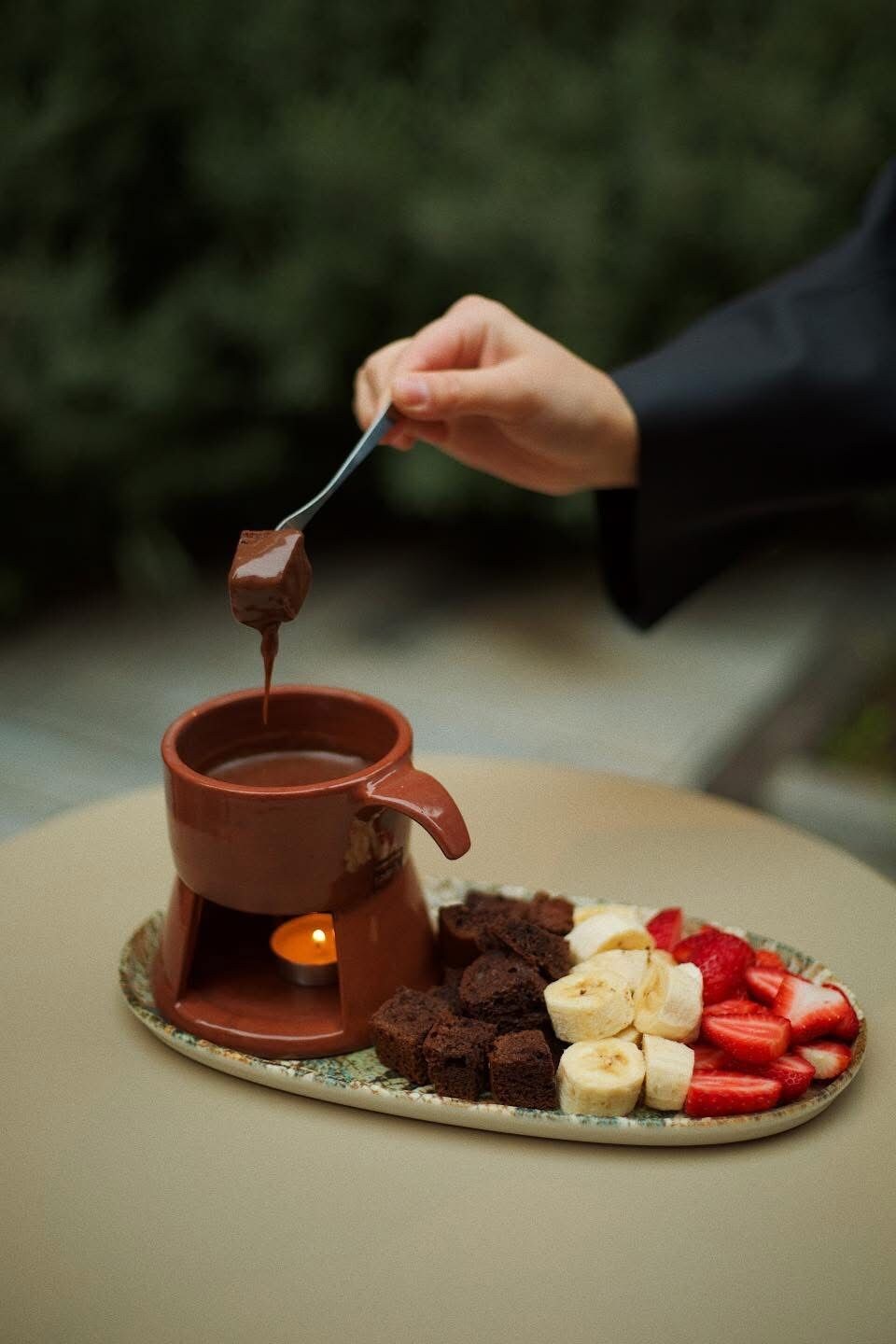 Una persona sumergiendo una cuchara cubierta de chocolate en una olla de chocolate caliente, con un plato de brownies, rodajas de plátano y fresas a un lado.