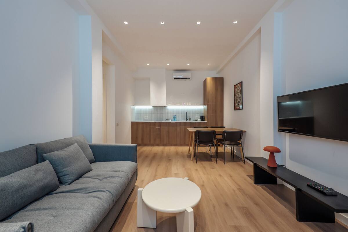 Ein modernes Wohnzimmer mit einer grauen Couch, einem kleinen weißen Tisch, einem Fernseher und einer Küche im Hintergrund.