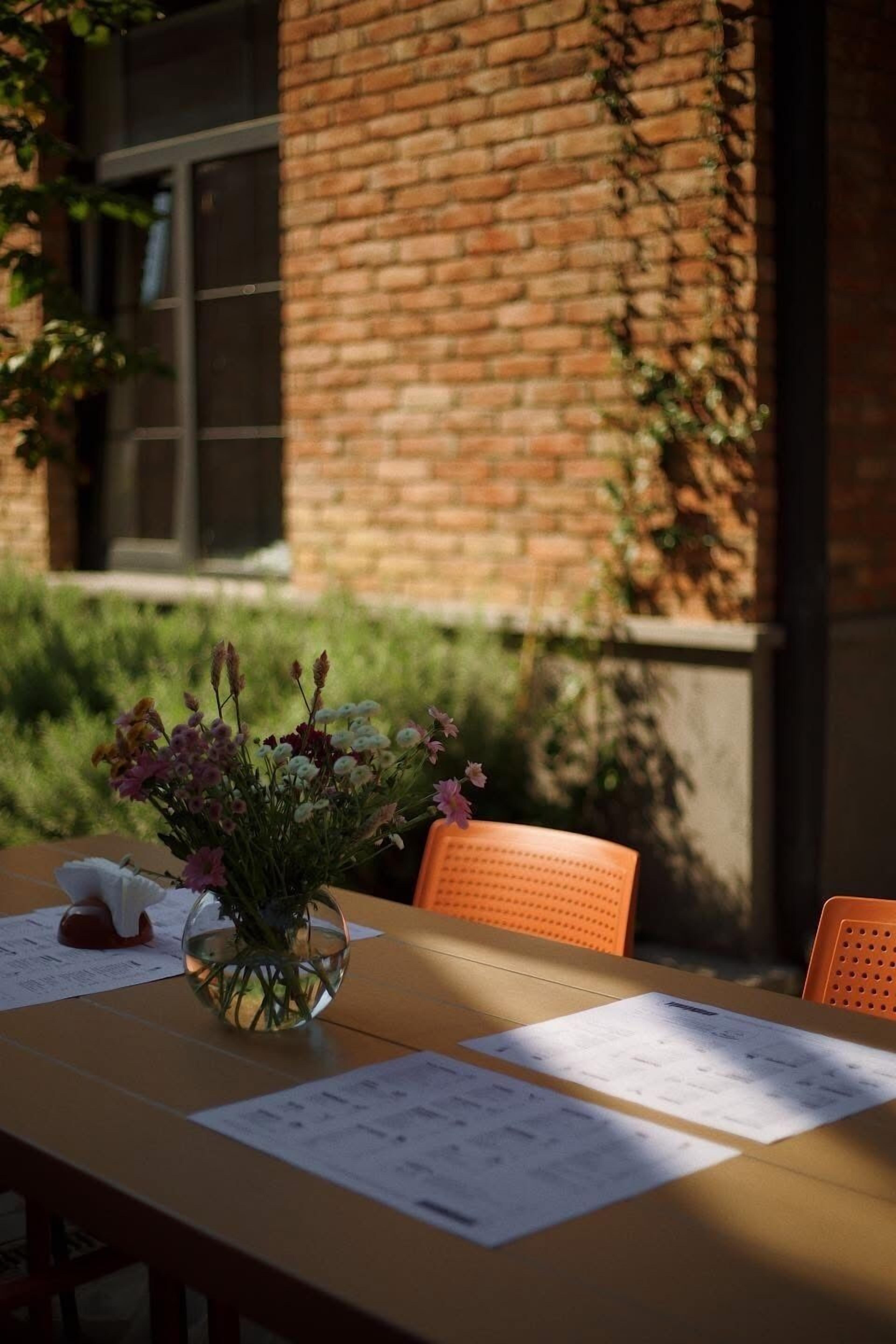 A table with papers, a glass vase with flowers, and a brick wall in the background.