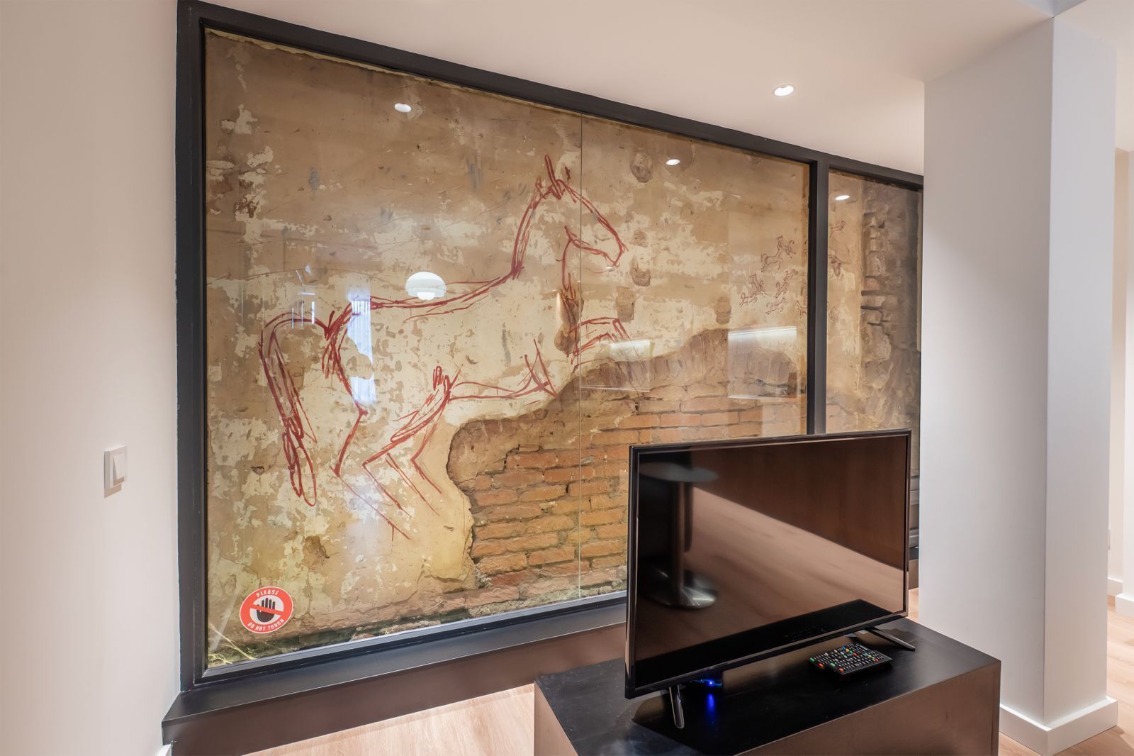 Una sala moderna con un cuadro abstracto grande en la pared, un televisor de pantalla plana sobre un soporte de madera, y una pared de ladrillo parcialmente visible detrás del cuadro.