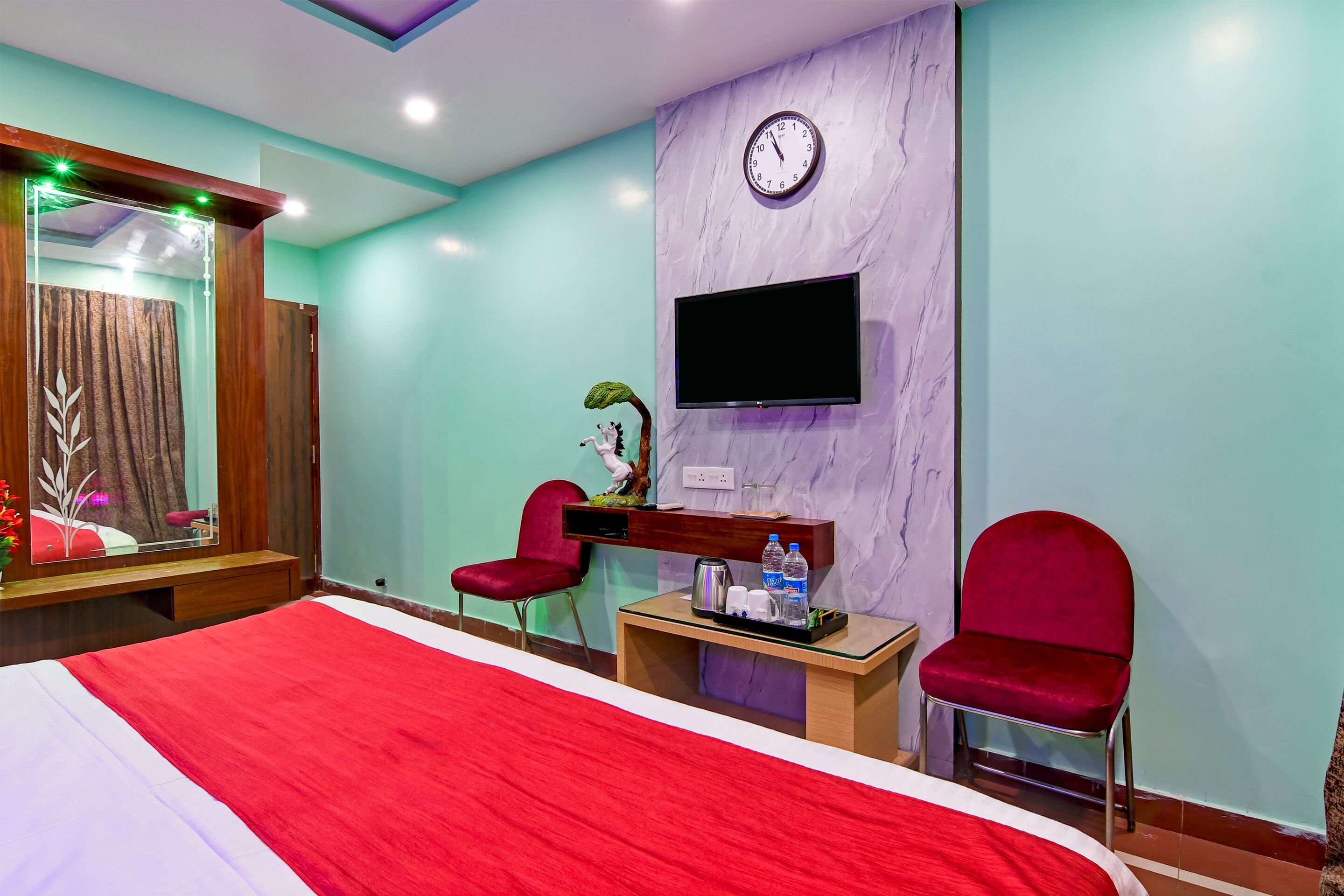 Habitación de hotel con una cama grande, dos sillas rojas, una mesita con un televisor y un reloj en la pared.