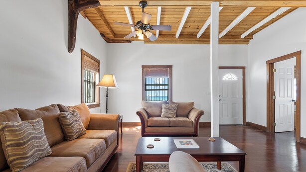 A spacious living room with a wooden ceiling, a ceiling fan, a brown leather sofa, a brown armchair, a coffee table, and a patterned rug. - Sunnyside Cottage (Cockburn Town)