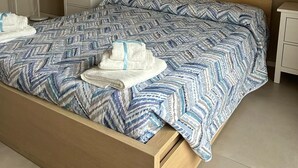Quarto com estrutura de cama de madeira, colcha estampada, obra de arte na parede, abajur e mesa de cabeceira.