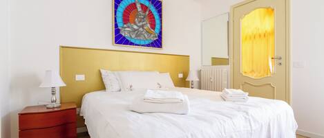 A neatly made bed with white linens, a wooden nightstand with a lamp, and a wall-mounted painting.