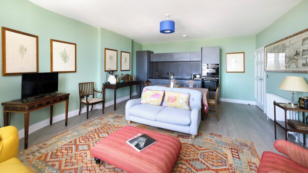 A living room with a sofa, ottoman, and chairs, a television, a kitchen area, and framed botanical prints on the walls. - Inviting Flat near City Centre (Edinburgh)