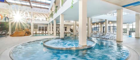 Une grande piscine intérieure à forme circulaire, entourée de chaises longues et dotée d’un plafond en verre qui laisse passer la lumière naturelle.