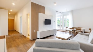 En moderne stue med et fladskærms-tv, grå sofaer, et spiseområde med træstole og et stort vindue med udsigt over grønne omgivelser.