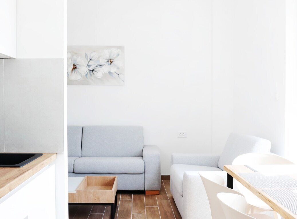 A modern living room with a grey sofa, a white armchair, and a wooden coffee table. A painting of white flowers hangs on the wall.