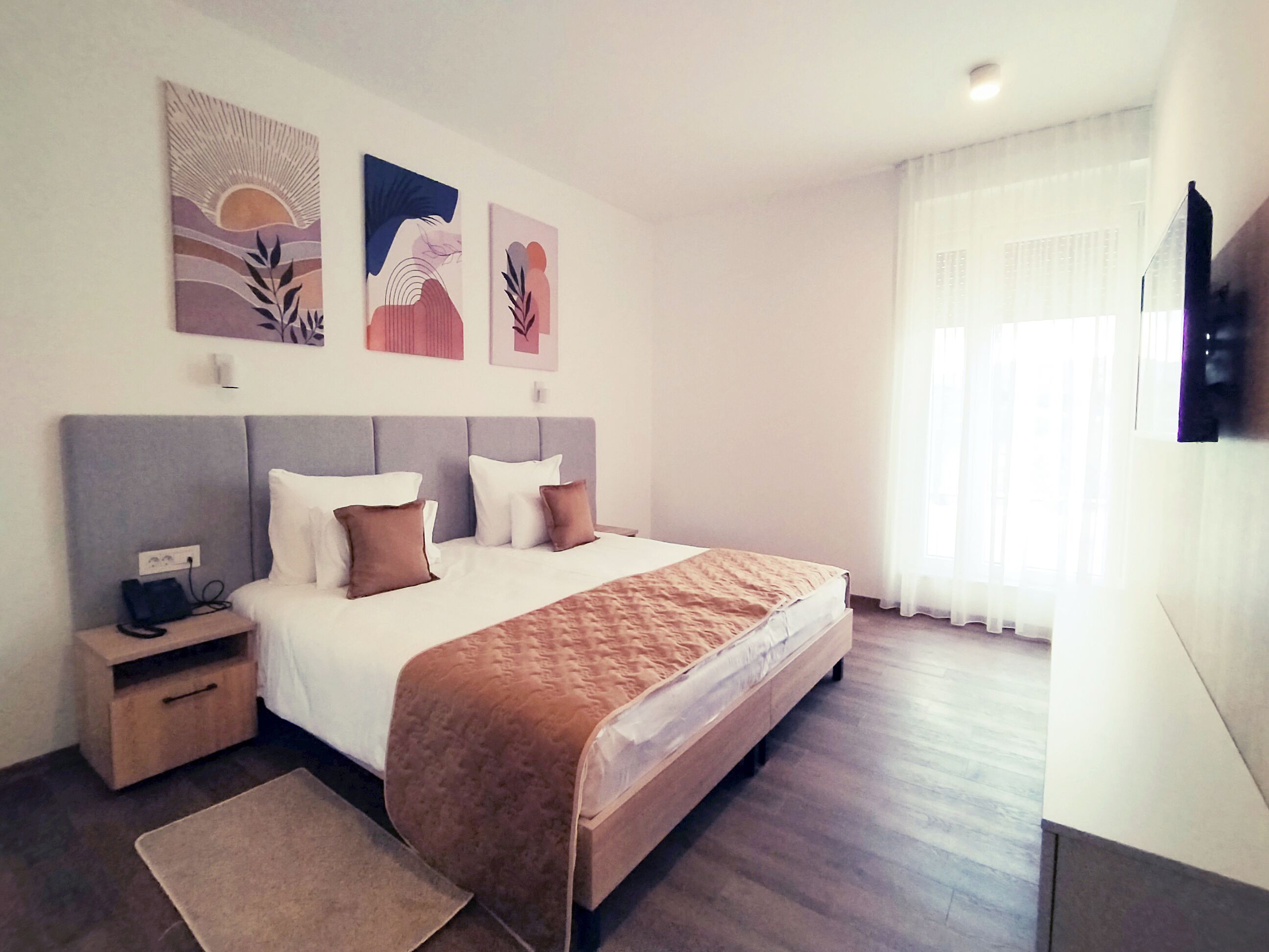 A modern hotel room with a large bed, a bedside table, a flat-screen TV mounted on the wall, and artwork above the bed.