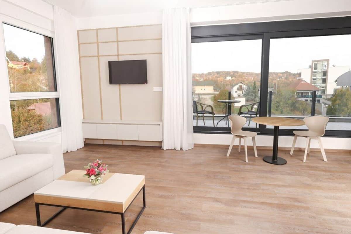 Una sala moderna con un ventanal grande, un televisor de pantalla plana, un sofá, una mesa de centro con flores y un balcón con sillas y una mesa.