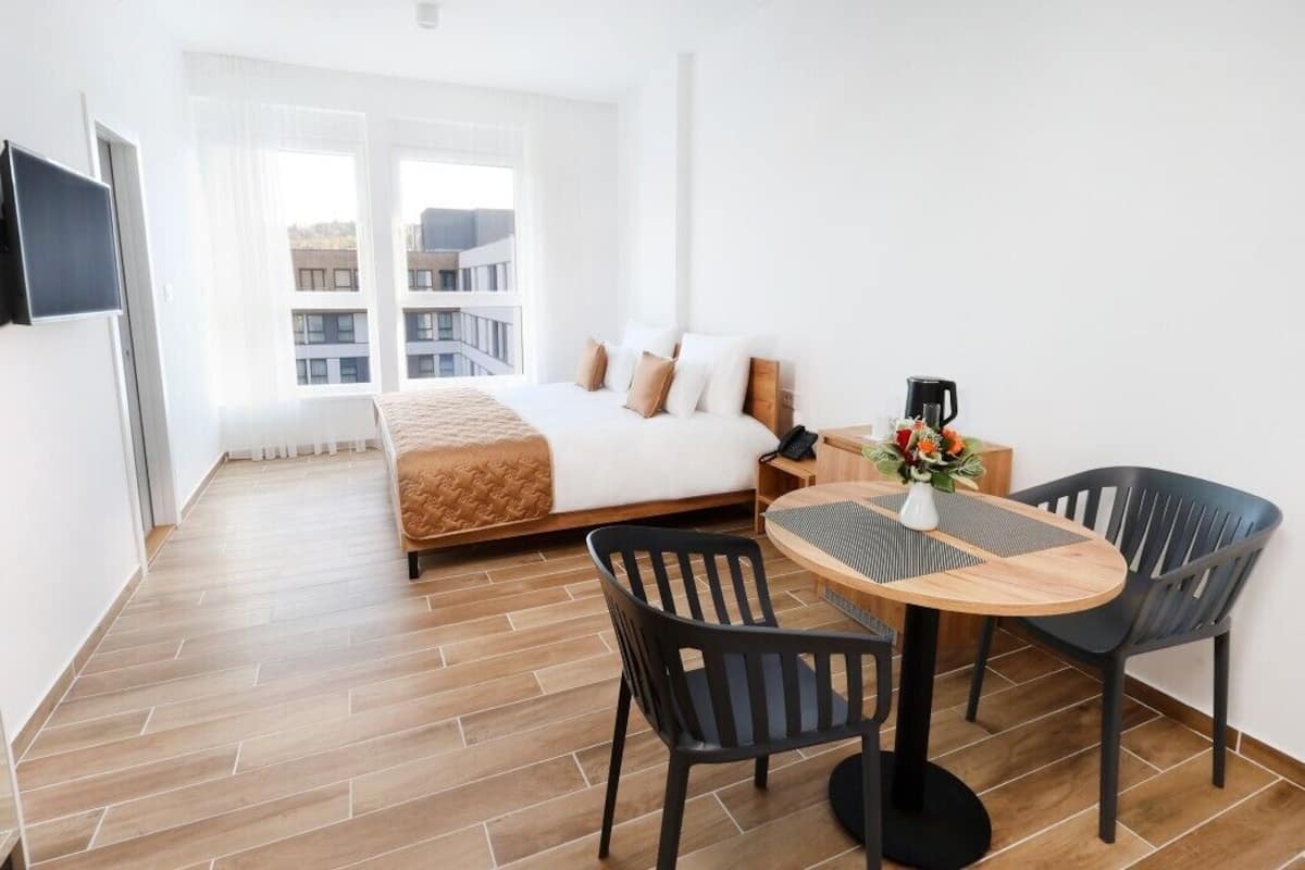 Habitación de hotel moderna con cama, una mesa de comedor con sillas, televisor y vistas a edificios a través de amplias ventanas.