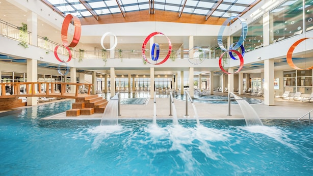 En indendørs swimmingpool med flere vandstråler, et glasloft og moderne arkitektur.