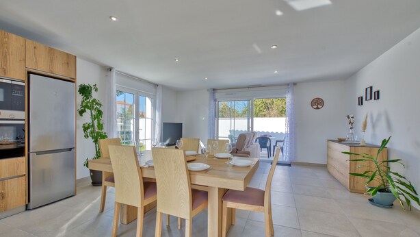 A modern kitchen with a built-in oven and refrigerator, a dining area with wooden chairs and a large table, and a view of the outdoors through large windows.