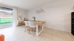 Un comedor con una mesa de madera, sillas blancas y vistas a un edificio verde en el exterior.