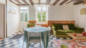 Un salon chaleureux avec un canapé vert, une table ronde entourée de chaises, un plafond en bois et une grande fenêtre.
