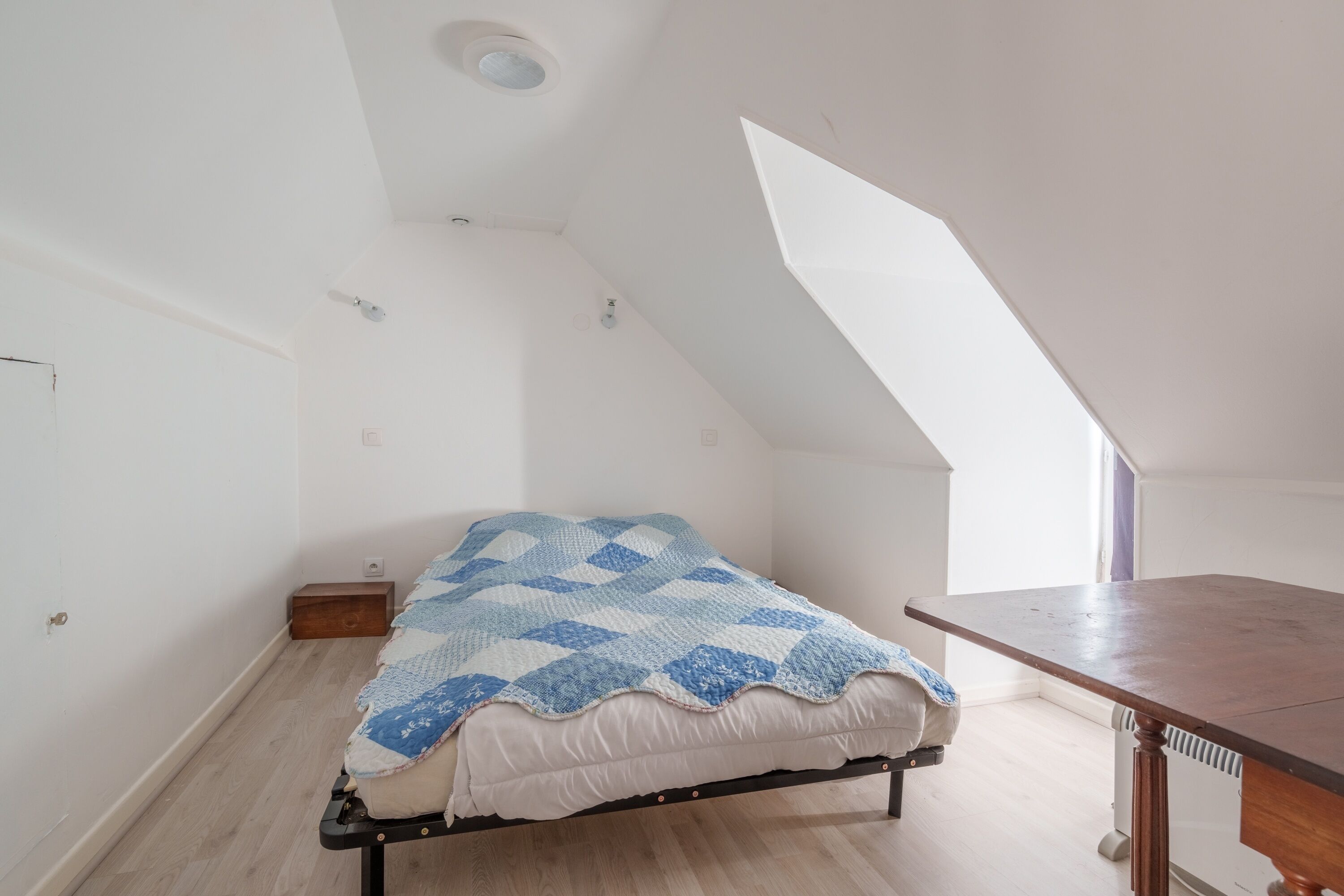 Una habitación pequeña con cama individual, techo inclinado y una mesa de madera.
