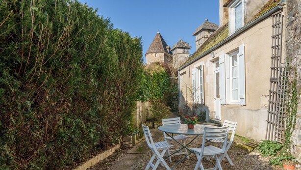 Une cour avec une table ronde et des chaises blanches, entourée d’un mur de pierre et d’une végétation luxuriante.