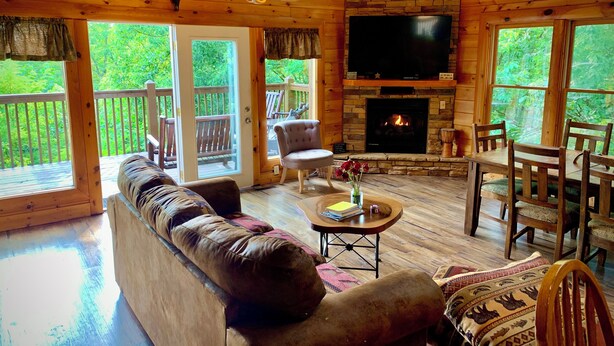Interior acogedor de una cabaña con chimenea de piedra, un televisor grande, techo de madera y vistas al exterior.