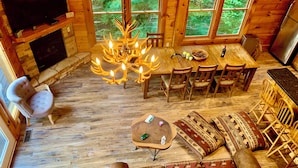 Interior acogedor de una cabaña con chimenea, un candelabro, una mesa de comedor con sillas, un conjunto de sofás y un televisor montado en la pared.
