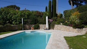 A rectangular swimming pool with a stone border, surrounded by a stone wall and lush greenery, including tall trees and flowering plants. - Beautiful house, very well located, sleeps 4 to 10, with swimming pool and air conditioning. (Saint-Raphaël)