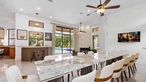 Un vaste espace dédié à la salle à manger, avec une grande table en marbre et des chaises à revêtement blanc, un ventilateur de plafond et une vue sur une piscine, visible à travers de grandes fenêtres.