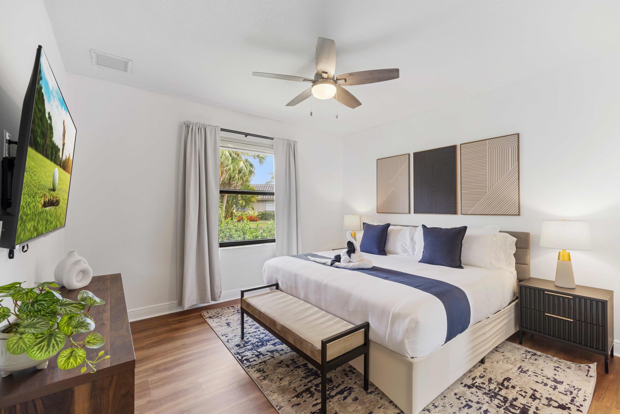 A modern hotel room with a large bed, a ceiling fan, a TV, a bedside table with a lamp, and a window with curtains.