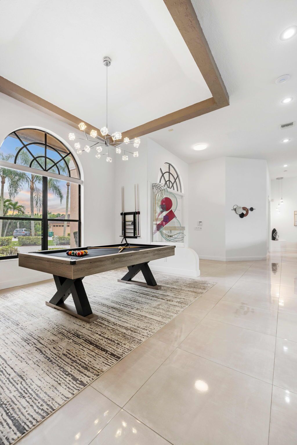 A modern indoor pool table with a wooden finish, a patterned rug, and a view of palm trees through a large arched window.