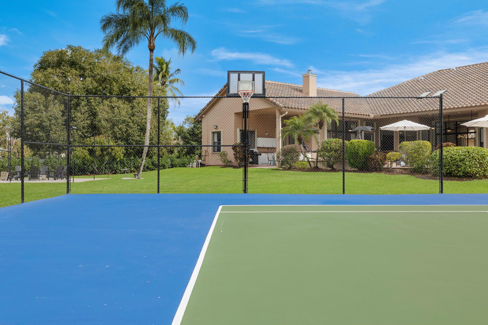 A tennis court with a basketball hoop, surrounded by a fence, palm trees, and a building with a tiled roof.