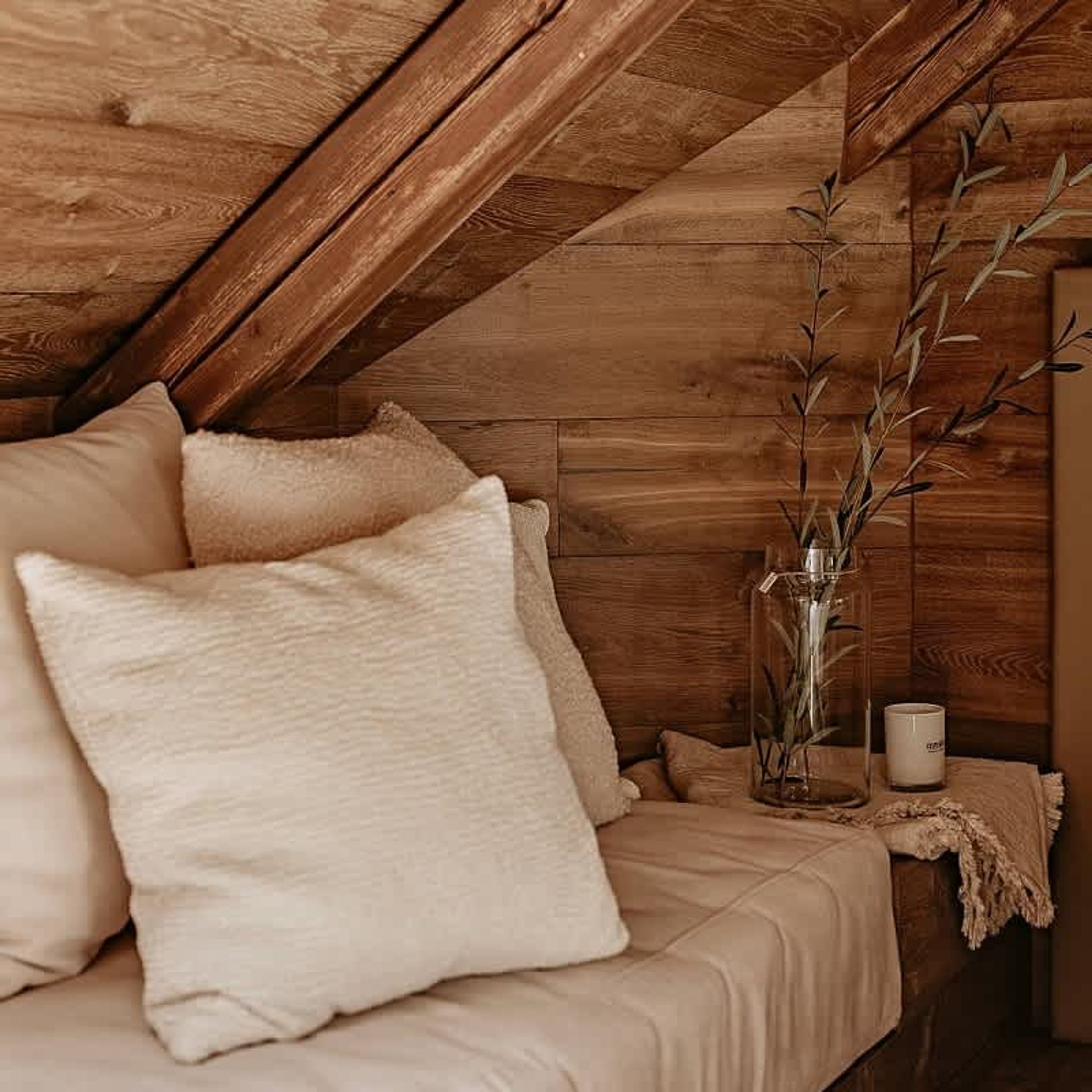 A cozy attic bedroom with wooden beams, a bed with beige bedding, a vase with branches, and a candle on a nightstand.