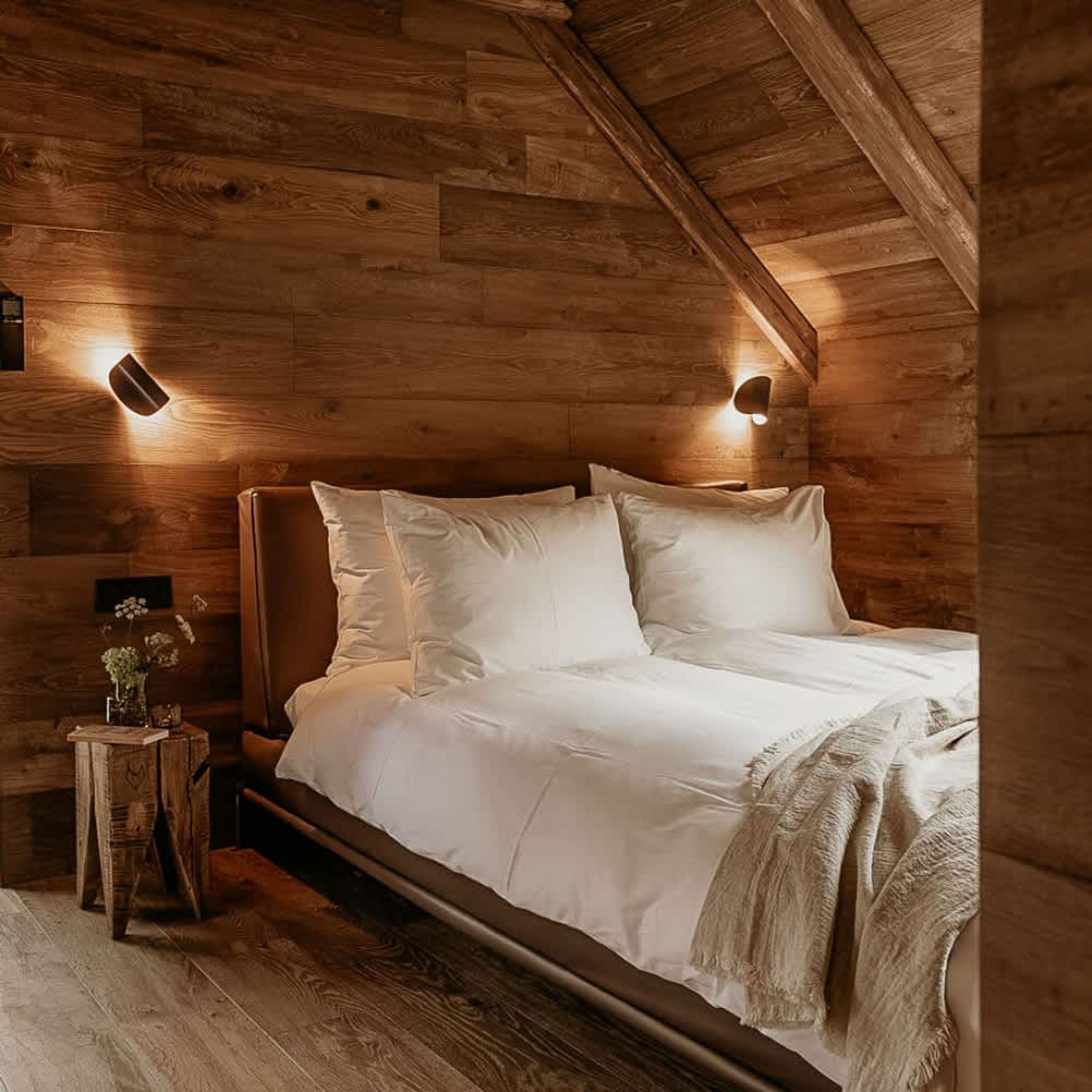 Quarto acolhedor de cabana de madeira, com uma cama, duas luminárias fixadas na parede e uma pequena mesa de cabeceira de madeira com um vaso.