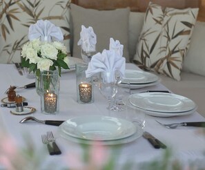 Una mesa de comedor dispuesta con platos blancos, cubiertos y cristalería, acompañada de flores blancas y velas.