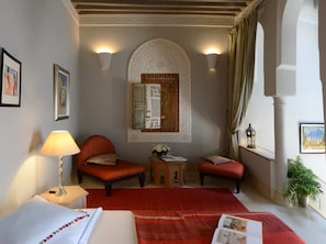 Habitación de hotel con alfombra roja, dos sillones rojos, una cama blanca, una mesita con una lámpara y un elemento decorativo en la pared.