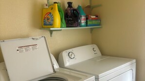 A front-loading white washer and dryer in a laundry room with a shelf above containing cleaning supplies.
