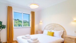 A well-lit bedroom with a large bed, a bedside table, a nightstand, and a view of a hillside through the window. - GuestReady - A sweet escape in Caniço (Caniço)