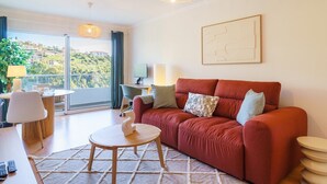 A modern living room with a red sofa, a round wooden coffee table, and a view of a balcony with a cityscape. - GuestReady - A sweet escape in Caniço (Caniço)